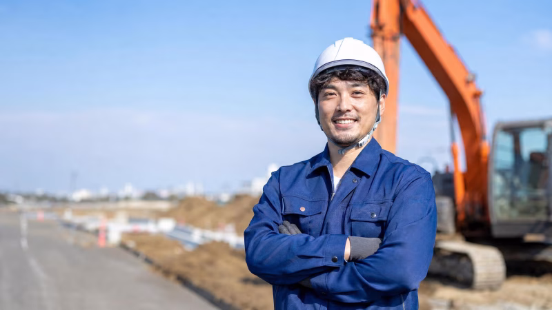 建設現場で腕を組む男性重機オペレーターと油圧ショベル